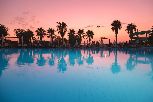 Beautiful reflection in swimming pool at colorful sunset. Purple sky reflected in water, palm trees, sun beds, umbrellas at night in summer. Luxury resort. Landscape with empty pool in twilight