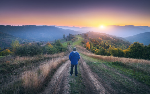Man on the dirt road on the hill and mountains in fog at colorful sunset in autumn in Ukraine. Landscape with guy, foggy hills, green grass, meadows, trail, forest, beautiful sky in fall at dusk