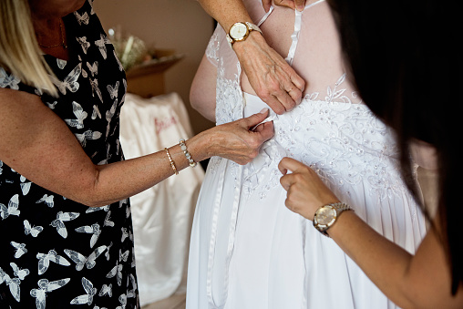 Body positive future bride getting dressed with mother and friend before LGBTQ+ wedding. They are lacing the back of the dress. No faces. This is part of a series about a lesbian couple getting married. Horizontal indoors close-up shot with copy space.