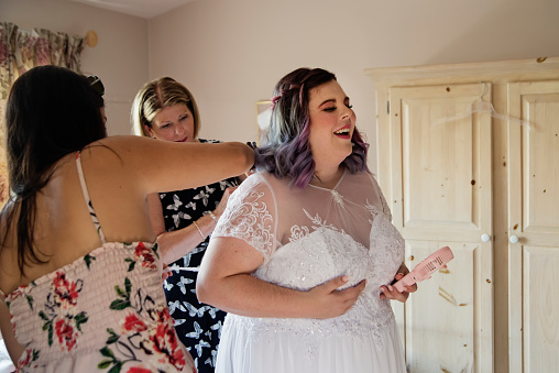 Body positive future bride getting dressed with mother and friend before LGBTQ+ wedding, while holding a small fan. This is part of a series about a lesbian couple getting married. Horizontal indoors waist up shot with copy space.