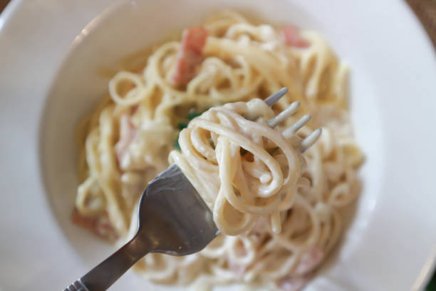 A hot, freshly prepared plate of creamy carbonara pasta with a rich, cheesy sauce. A person is twirling spaghetti with a fork while steam rises from the dish, highlighting its freshness.
