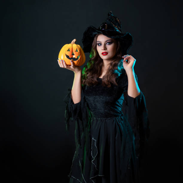 A young beautiful witch in a conical hat holds a Halloween pumpkin in her hands and makes a spell with her hand. Halloween Costume Party, Witches' Sabbath
