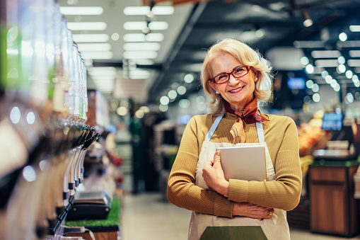 Portrait of happy senior woman holding digital tablet, standing between shelves with food and enjoying her job