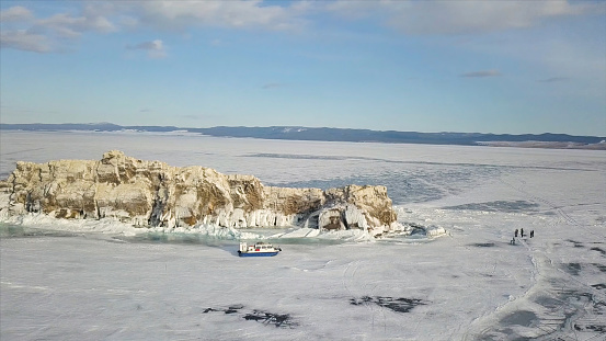 People travel on the ice of frozen Baikal Lake near safe and comfortable Khivus hovercraft. Aerial view of a group of tourists walking on thick ice of amazing lake, extreme tourism concept.