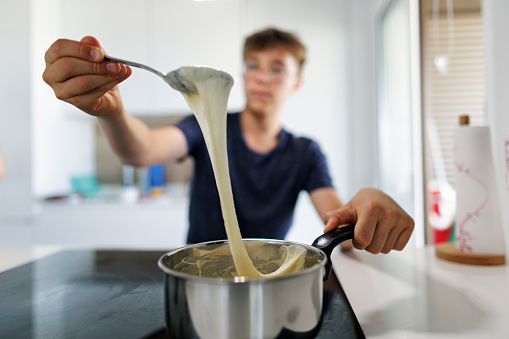 Teenage boy is helping to prepare lunch for the family. Boy is cooking fondue in the pot on the stove.\nHe is raising a spoon with delicious gooey, molten cheese.\nShot with Canon R5