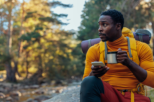 A young African American hiker is sitting on a rock in the mountains drinking water from his cup and checking online maps on his smartphone.