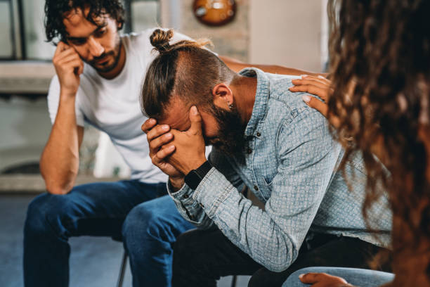 People are comforting a man during a group therapy session People are comforting a man during a group therapy session. Mental health concept in a loft with big windows. Healthcare and medicine concept. Group of people sitting in a circle are participating in a support meeting, talking with each other. man crying stock pictures, royalty-free photos & images