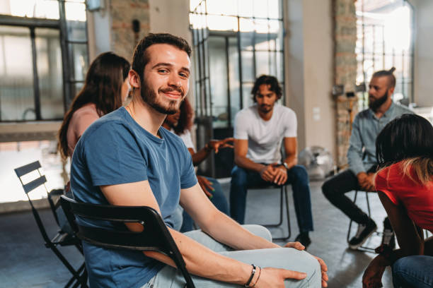 Portrait of a man looking at camera during a group therapy session Portrait of a man looking at camera during a group therapy session. Mental health concept in a loft with big windows. Healthcare and medicine concept. Group of people sitting in a circle are participating in a support meeting. junkie stock pictures, royalty-free photos & images