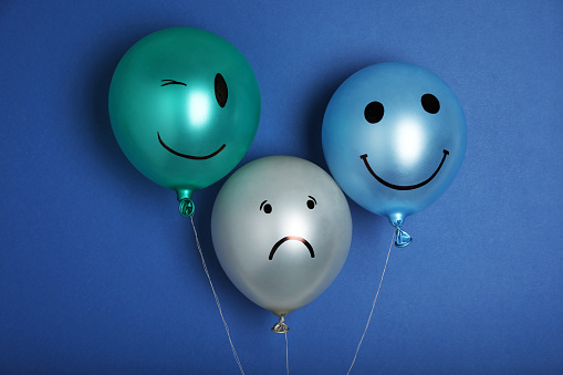 Balloon with sad face among happy ones on blue background. Depression concept