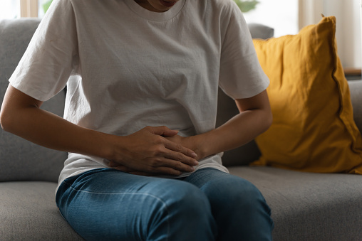 Close-up unrecognize woman suffering from stomachache or menstruation pain. Unhealthy teenage girl sitting on sofa, touching her belly feeling pain and unwell.