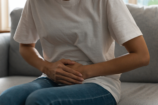Close-up unrecognize woman suffering from stomachache or menstruation pain. Unhealthy teenage girl sitting on sofa, touching her belly feeling pain and unwell.