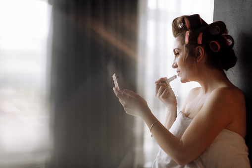 beautiful young woman with curlers is doing make-up while looking at the mirror on bed in the morning.