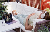 Adult senior plus size woman having online hypnotherapy session with psychotherapist lying on sofa on outdoor terrace at home.