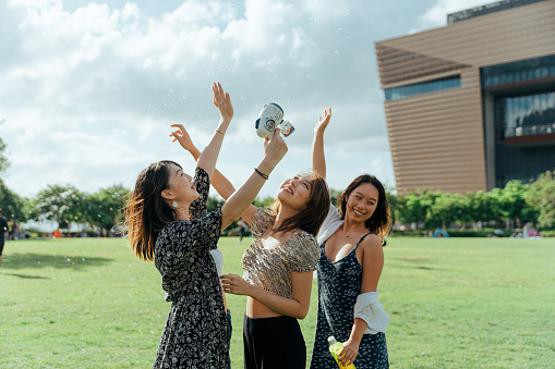 Three young Asian women hanging out together in a public park and playing with the bubble gun, while looking at the bright blue sky. A group of happy best friends playing and blowing bubbles with the bubble machine under sunlight on the grass field in a park, smiling towards the view of the sky. Girls enjoying a sunny day outdoors and having fun together.