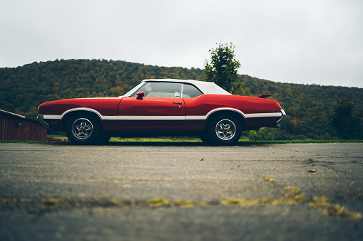 From below of red old vintage American car parked in countryside against gray overcast sky and green hill on background