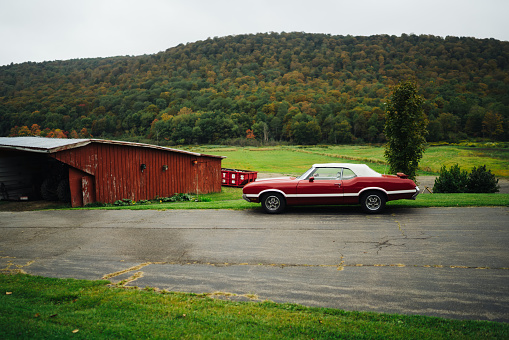 Side view of red retro American car parked near shabby barn in countryside against gray overcast sky and green hill