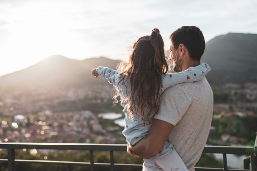 Young father and daughter in mountains at sunset looking at the city.