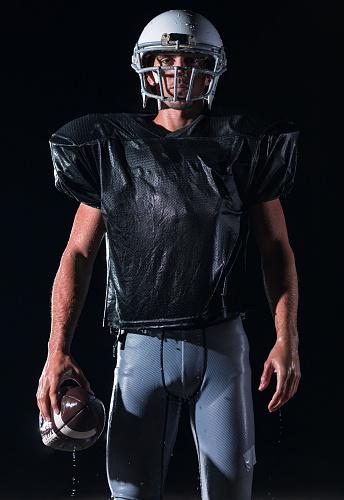 American Football Athlete Warrior Standing on a Field Holds his Helmet and Ready to Play. Player Preparing to Run, Attack and Score Touchdown. Rainy Night with Dramatic lens flare and rain drops. High