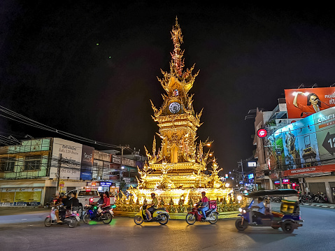 Chiang Rai, Thailand - September 16, 2019: taxis motorcycles and tuktuk passing in front of Thai style gold clock tower in Chiang Rai illuminated at night