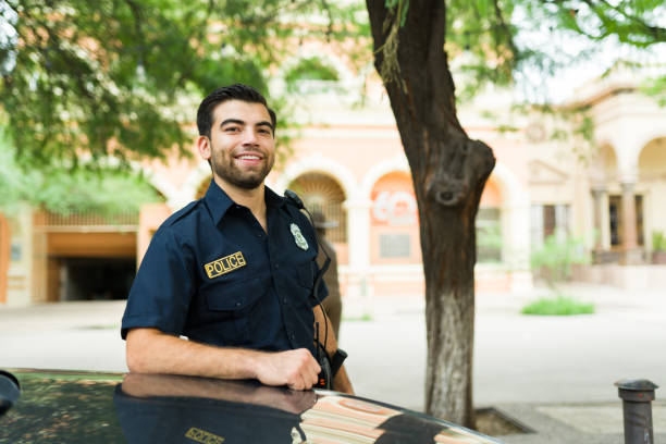 Portrait of a happy male cop leaning into the police car Attractive young man in a uniform smiling looking at the camera while working as a police officer in the city streets police force smiling officer stock pictures, royalty-free photos & images