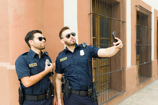 Attractive police agents on duty smiling and looking happy outdoors taking a selfie with a smartphone