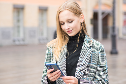 A young Caucasian blonde girl in a coat uses a mobile phone on the street in the city on an autumn day. A satisfied young girl is working from a mobile phone online on the Internet.
