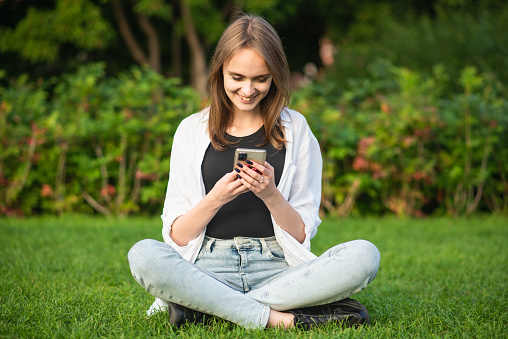 A young and attractive Caucasian girl in casual clothes is sitting on the grass in the park and using a mobile phone.