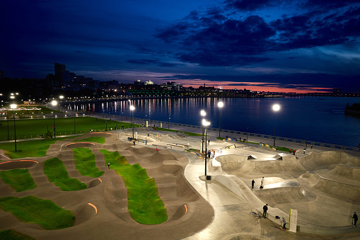 Evening view of the city embankment. Rare unrecognizable people in the illuminated skatepark. Blue sky and river, green grass. Above view. Kazan city, Russia