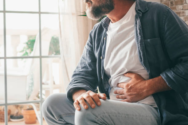man suffering with severe stomach pain sitting at home. hand of mature guy holding abdomen suffering from ache, diarrhea or indigestive problem. caucasian guy pressing on belly on painful sensation - buik stockfoto's en -beelden