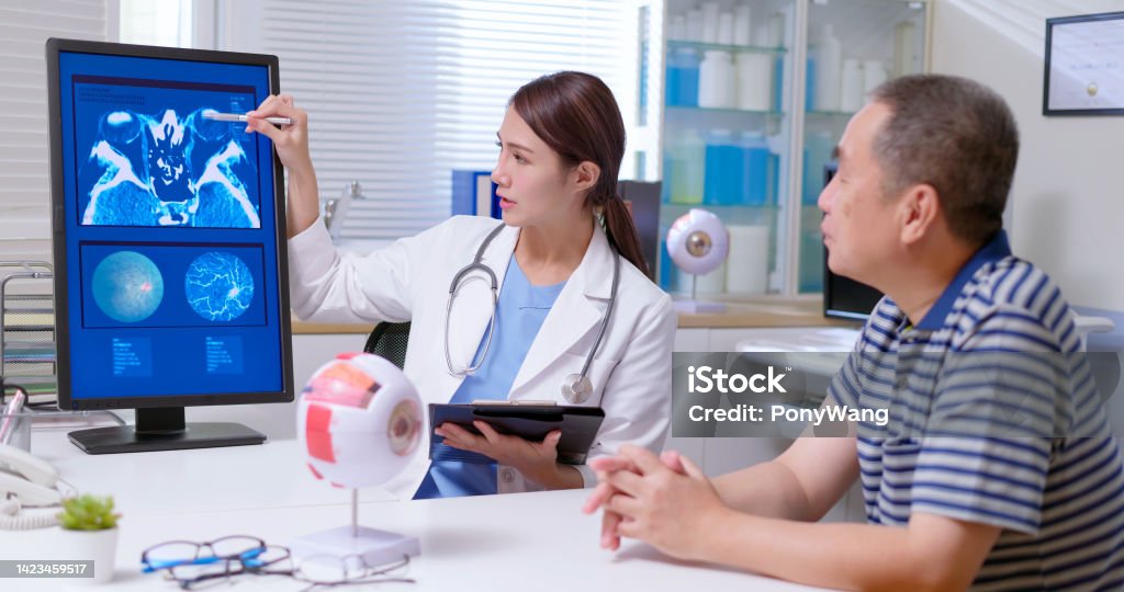 doctor explain eye xray film attractive young brunette ponytail female asian doctor wear white coat and stethoscope pen pointing explaining xray film on computer to gray hair old sick man at clinic - an eye model display Eye Stock Photo