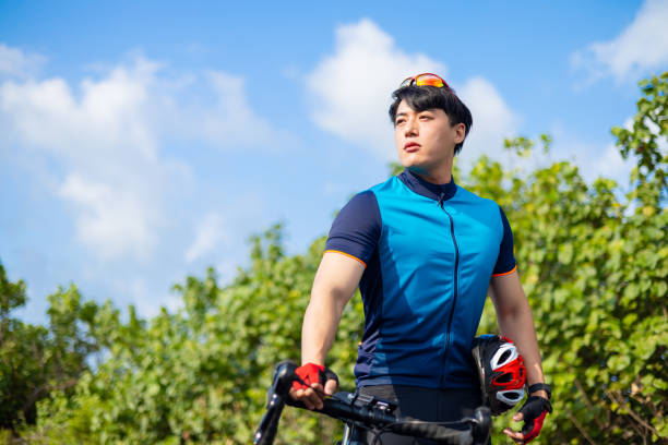 asian young male cyclist with helmet in hand standing by his bike on the road is looking far away