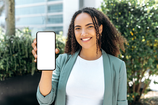 Confident lovely happy young mixed race woman, with curly hair, in stylish formal wear, stands outdoors, holds cellphone in hand, shows a white blank mock-up screen, space for your advertising, smile