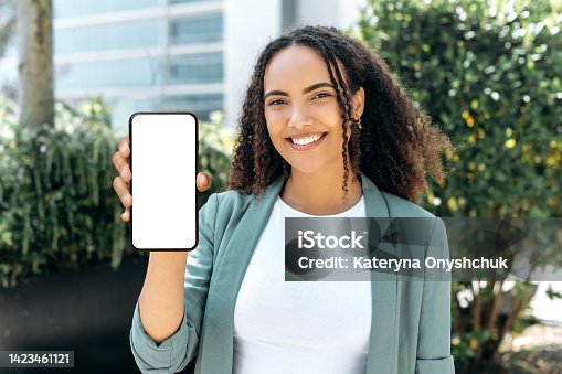 istock Confident lovely happy young mixed race woman, with curly hair, in stylish formal wear, stands outdoors, holds cellphone in hand, shows a white blank mock-up screen, space for your advertising, smile 1423461121