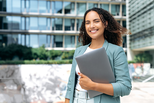 Portrait of confident successful young mixed race curly woman, formally dressed, business woman standing with laptop outdoors against the background of the business center, looks at camera, smiling
