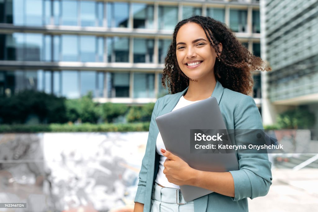 Portrait of confident successful young mixed race curly woman, formally dressed, business woman standing with laptop outdoors against the background of the business center, looks at camera, smiling Manager Stock Photo