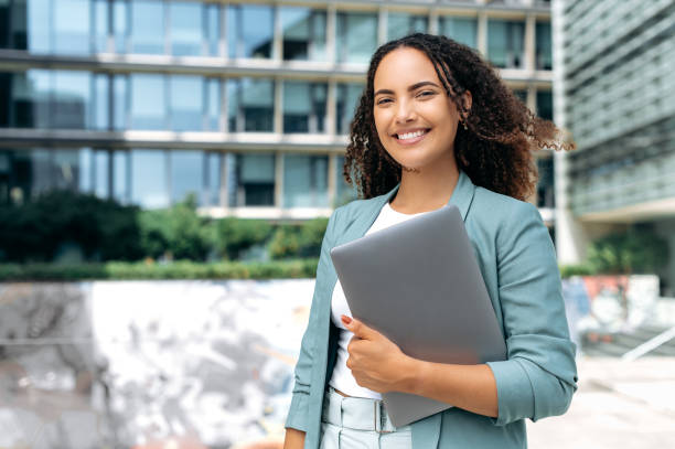 Portrait of confident successful young mixed race curly woman, formally dressed, business woman standing with laptop outdoors against the background of the business center, looks at camera, smiling Portrait of confident successful young mixed race curly woman, formally dressed, business woman standing with laptop outdoors against the background of the business center, looks at camera, smiling secretary stock pictures, royalty-free photos & images