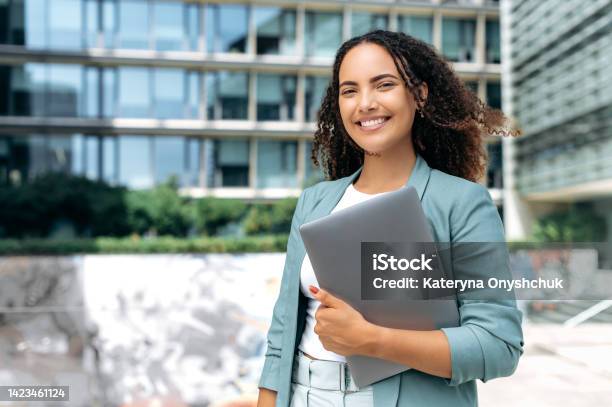 Portrait Of Confident Successful Young Mixed Race Curly Woman Formally Dressed Business Woman Standing With Laptop Outdoors Against The Background Of The Business Center Looks At Camera Smiling Stock Photo - Download Image Now