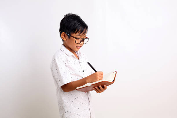 istock Smart asian schoolboy standing while writing on the book. Isolated on white background with copyspace 1423481286