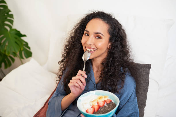 A beautiful multi-ethnic woman eats fruit bowl in bed licking the spoon A beautiful multi-ethnic woman eats fruit bowl in bed. High quality photo eating chia seeds stock pictures, royalty-free photos & images