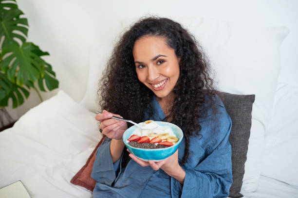 a beautiful multi-ethnic woman eats fruit bowl in bed - multi well trays imagens e fotografias de stock