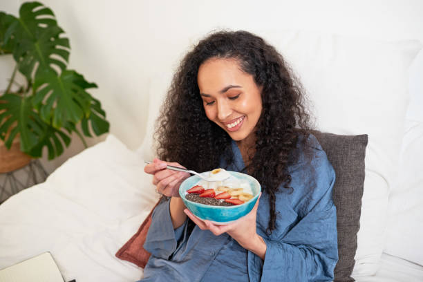 a beautiful multi-ethnic woman eats fruit bowl in bed - multi well trays imagens e fotografias de stock