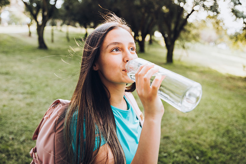 Young teenage girl drinking water from a glass bottle in the public park. Environmental conservation.
