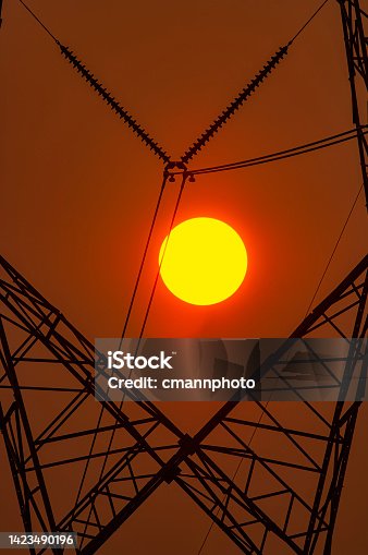 istock Close-up of the sun setting behind a high voltage electric transmission tower on a hot summer day 1423490196