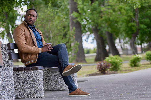 Handsome black man sits on a bench in the park waiting for another person. An African-American man sat down on a bench in an alley in a park, resting from a long road.