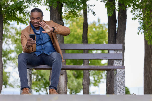 ハンサムな黒人ブロガーは、木製のベンチに座りながら現代の携帯電話を使用しています - correspondence waving horizontal outdoors ストックフォトと画像