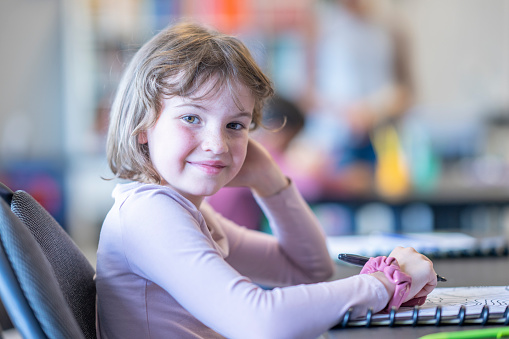 A sweet little Montessori student sits at her desk as she works away independently on an assignment.  She is dressed casually, has a notebook open in front of her and a pencil in hand as she looks up from her work to smile for the camera.