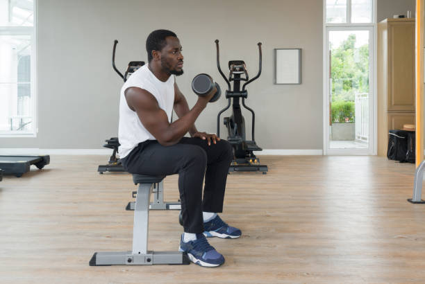 istock Young short curly black hair man with moustache and beard working on his biceps, lifting barbell with one hand on Multi-Function Bench. There are cardio machines in the gym. 1423495416