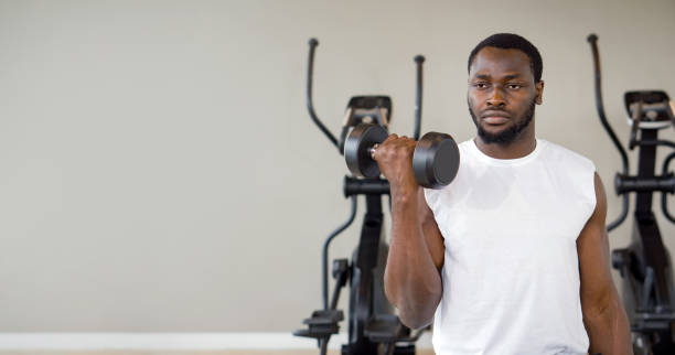 istock Young short curly black hair man with moustache and beard working on his biceps, lifting barbell with one hand. There are cardio machines in the gym. 1423495418