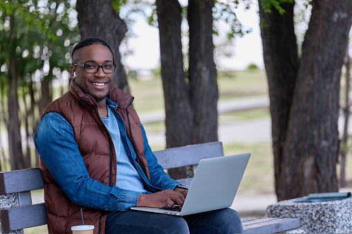Cheerful man in glasses and a brown vest sits on a bench with a laptop drinking coffee. A popular black blogger reports on the street in a park on a bench in clear weather