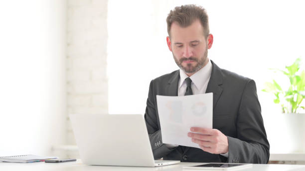 istock Young Businessman with Laptop Reading Documents in Office 1423499086